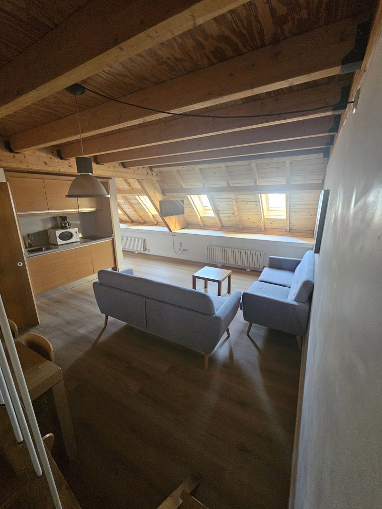 Sala de estar con techo y suelos de madera, mueble auxiliar, sofás y mesita. Al fondo tres ventanas aboardilladas