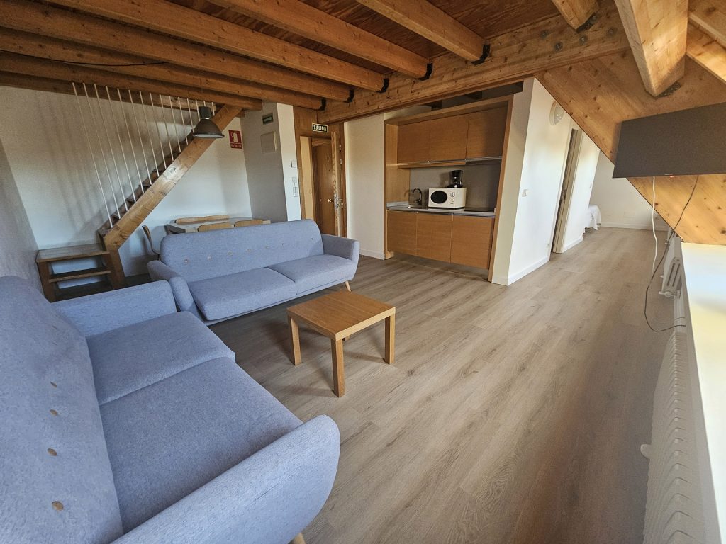 Sala de estar con techo y suelos de madera, mueble auxiliar, sofás y mesita. Al fondo la escalera