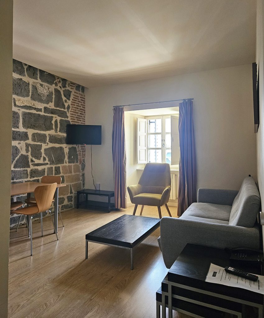 Vista de la sala, al fondo una ventana y una pared de piedra. Incluye dos sofás, dos mesitas auxiliares, mesa de comedor y butacas