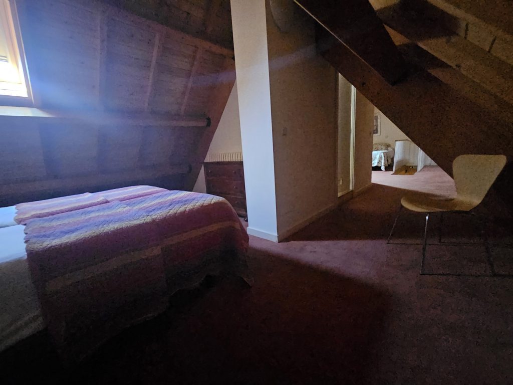 Vista de conjunto, en primer plano una cama, techo en madera aboardillado a dos aguas